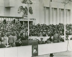 The image shows President Franklin Roosevelt delivering a speech on the opening day of the 1939 New York World's Fair. Standing at a podium adorned with the fair's emblem, he addresses the crowd with passion and conviction. The backdrop of the stage is adorned with the American flag and the slogan "Building the World of Tomorrow". The audience is a sea of people, and the scene is full of energy and excitement. The ALT description for this image is: "President Franklin Roosevelt delivers a speech at the opening day of the 1939 New York World's Fair, standing at a podium adorned with the fair's emblem, surrounded by the American flag and a large crowd of people.